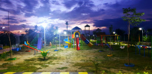 A brightly lit children's playground at dusk, featuring slides, swings, and a seesaw, set against a purple-hued sky with scattered clouds. The area is surrounded by trees and plants, creating a peaceful and inviting atmosphere.