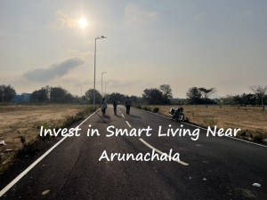 Three people walk along a paved road lined with streetlights, where a motorcycle is parked to the side. The sun shines through a partly cloudy sky. Overlaid text reads: ‘Invest in Smart Living Near Arunachala.