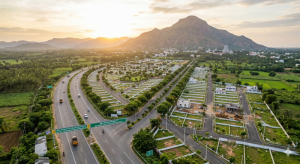 Aerial drone view of Tiruvannamalai highway corridor with plotted residential layouts, wide tar roads, green avenue trees, sunrise lighting, Arunachala hill in the distance, developing residential plots, photorealistic, 4K, peaceful South Indian town landscape